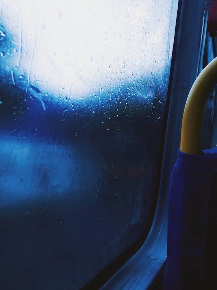 Services-03 Raindrops on a bus window create a moody, abstract scene with blurred blue hues.